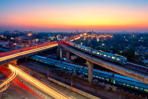 Aerial view of a city with elevated train tracks at sunset. A train, powered by Infrastructure Intelligence from Bentley Systems, travels on the bridge while light trails from cars are visible below. Buildings and a colorful sky create a stunning backdrop.