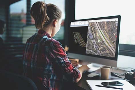 A person works on a computer displaying aerial maps in a modern office, focusing on infrastructure planning. A coffee cup and smartphone are on the desk, showcasing their dedication to excellence.