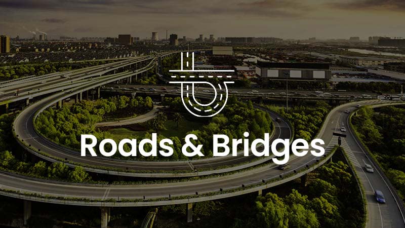 Aerial view of an intricate highway interchange with greenery surrounding it. "Roads & Bridges" text and logo are overlayed on the image. Cityscape and buildings are visible in the background.