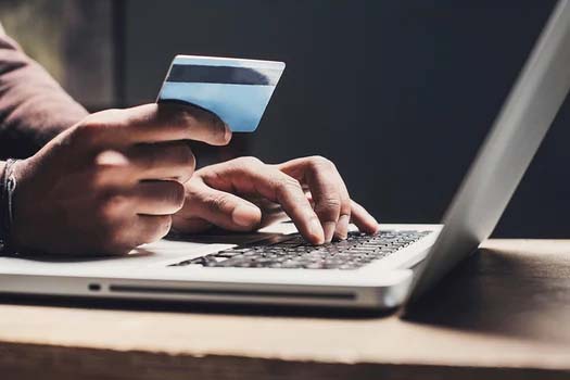 A person holds a credit card in one hand while typing on a laptop with the other hand, likely researching SEO strategies. The laptop sits on a wooden table.