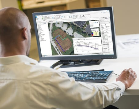 A person uses mapping software on a computer, displaying an overhead terrain view and various project details including water infrastructure, with one hand on the mouse and another on the desk.