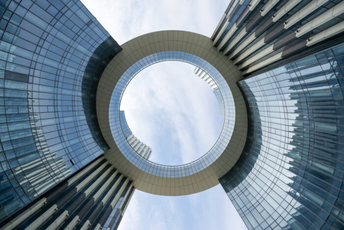 Upward view of two modern glass buildings connected by a large circular skybridge with an open center, reflecting the innovation of open infrastructure, set against a partly cloudy sky.