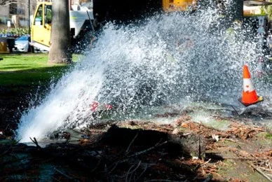 Water gushes from a burst pipe onto a city street next to an orange traffic cone, with a utility vehicle in the background, illustrating a situation that might require reference to the SewerGEMS