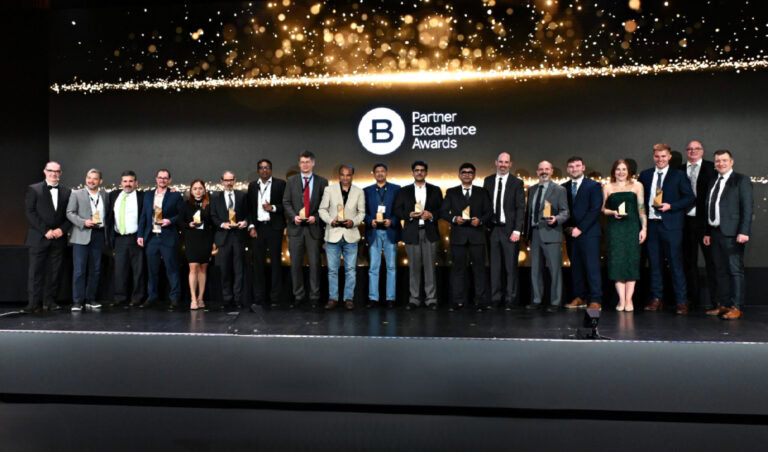 A group of people in formal attire stand on stage holding trophies at the Partner Excellence Awards, hosted by Bentley Systems, with a logo and gold lights displayed behind them.