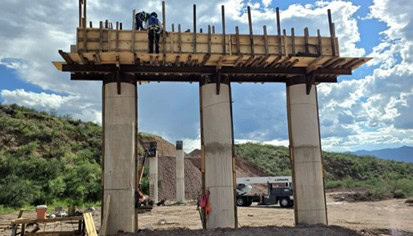 Three concrete bridge columns support a wooden formwork platform, with construction workers present; trucks and hilly terrain appear in the background under a partly cloudy sky for this Partner Spotlight project.