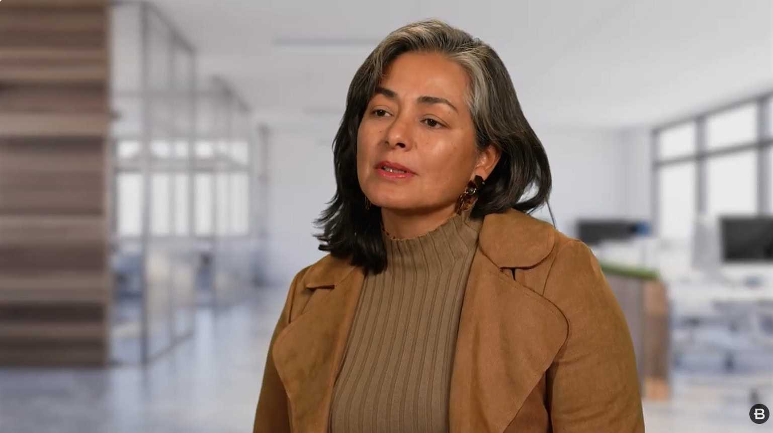 A woman with shoulder-length dark hair and gray streaks, featured in our Partner Spotlight, wears a tan jacket over a ribbed turtleneck sweater, standing in a modern office setting.