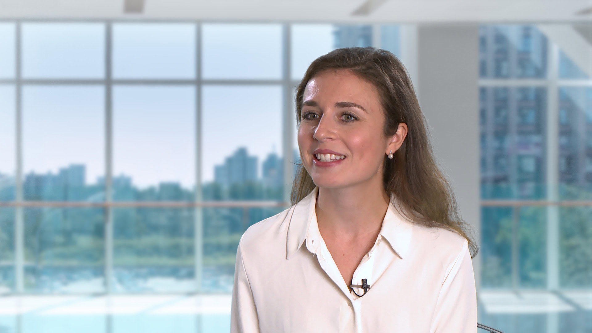 A woman with long brown hair wearing a white blouse sits indoors in front of large windows with a cityscape in the background.