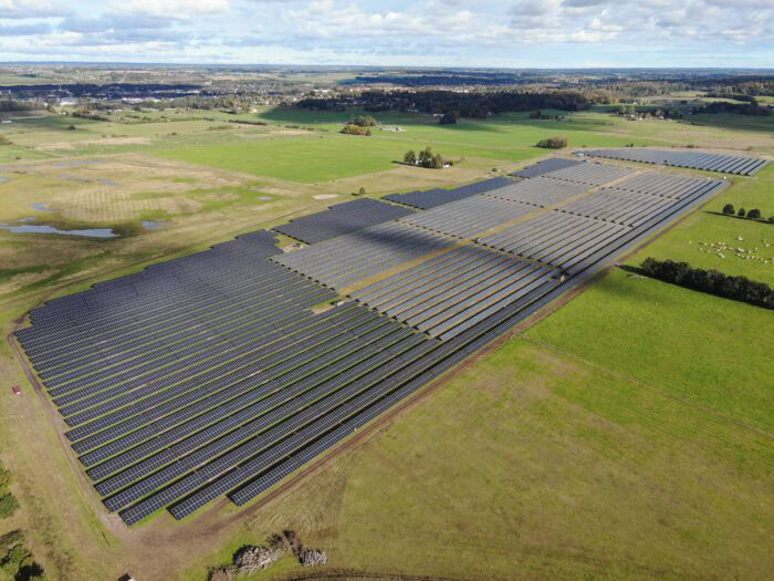 A large solar farm with rows of solar panels, installed across an open grassy field under a partly cloudy sky.