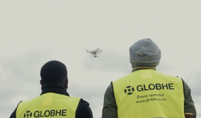 Two people in yellow "GLOBHE Drone operator" vests watch a drone soar through the cloudy sky.