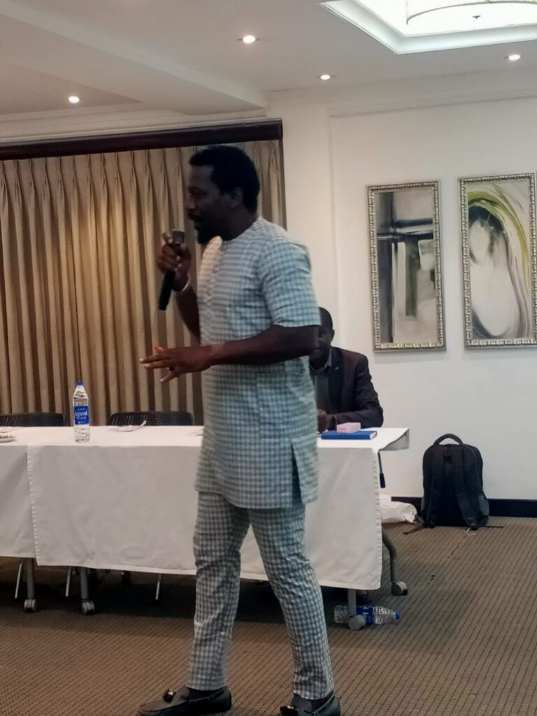 A man in a checked outfit speaks into a microphone in a conference room, sharing Future Concepts. A table with chairs, bottles, and framed artwork are visible in the background.