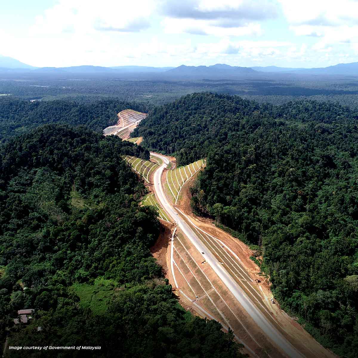 An award-winning, newly constructed highway cuts through a dense, green forest with mountains visible in the background under a partly cloudy sky.