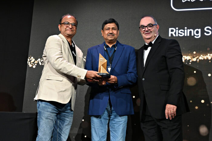 Three men in formal attire stand together on stage, with two of them holding an award. A backdrop behind them displays the words "Rising Star Winner"