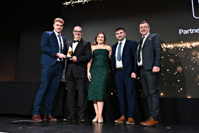 Five people in formal attire stand on stage, with one holding a trophy. A black background with gold accents is behind them, suggesting an awards event. | Bentley Systems | Infrastructure Engineering Software Company Five people in formal attire stand on stage, with one holding a trophy. A black background with gold accents is behind them, suggesting an awards event.