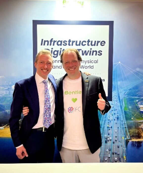 Two men in suits standing next to each other in front of a sign at Conference. | Bentley Systems | Infrastructure Engineering Software Company Two men in suits standing next to each other in front of a sign at Conference.