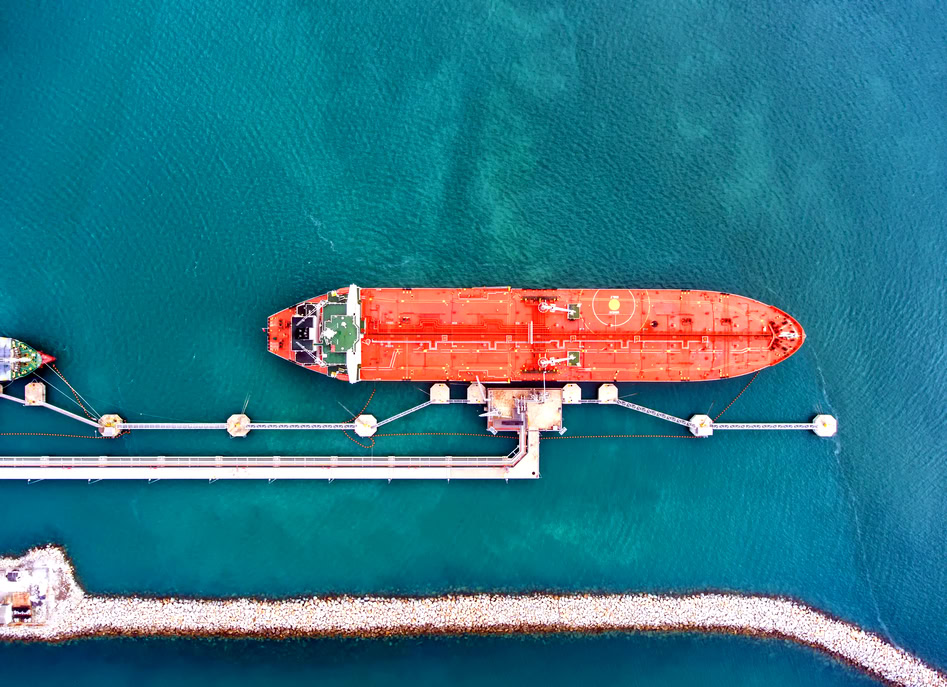 Aerial view of a large red cargo ship docked at a pier extending into blue-green water, with barriers around the docked area, highlighting the thoughtful design often seen in modern ports.