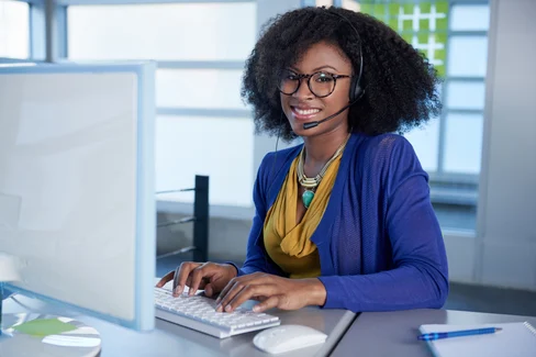 A woman wearing glasses and a headset is smiling at her desk, typing on a keyboard in an office setting, while watching an engaging OpenFlows video series.