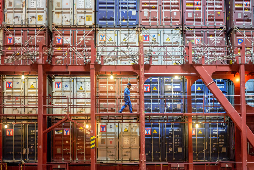 A worker in blue coveralls walks along a walkway in front of stacked shipping containers on a cargo ship, overseeing operations at busy ports.
