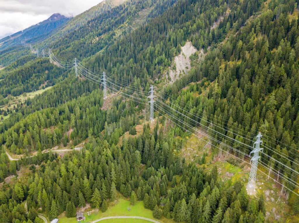 A series of high-voltage power lines and pylons run through a dense, green forested mountain landscape.