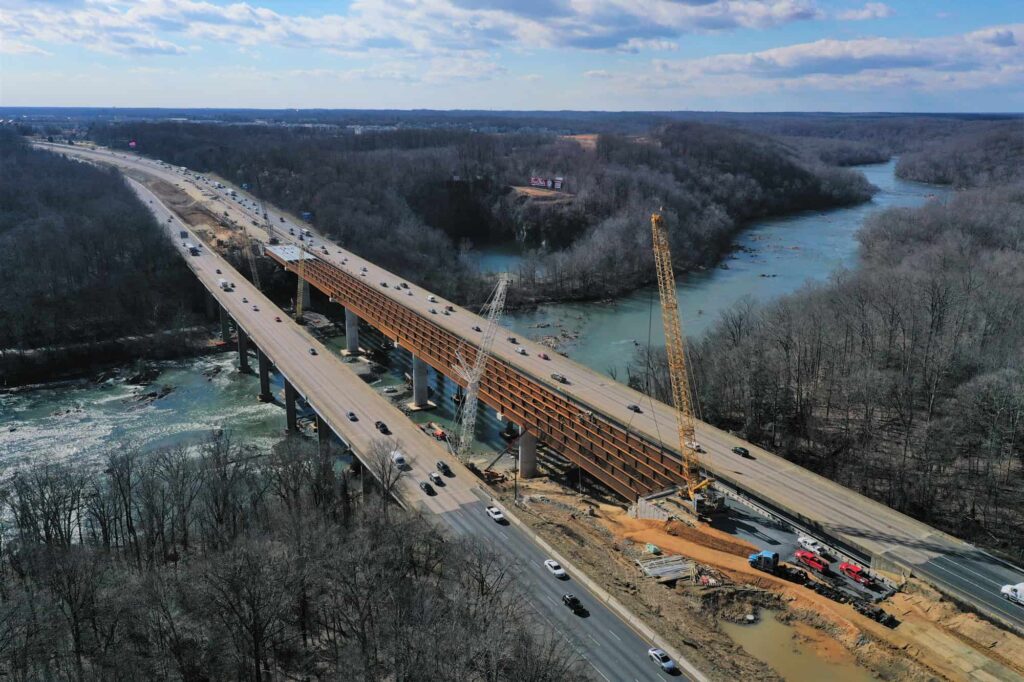 Aerial view of the Interstate Bridge project under construction over a river, alongside an adjacent finished bridge with vehicles. Cranes and construction equipment highlight progress on both structures.