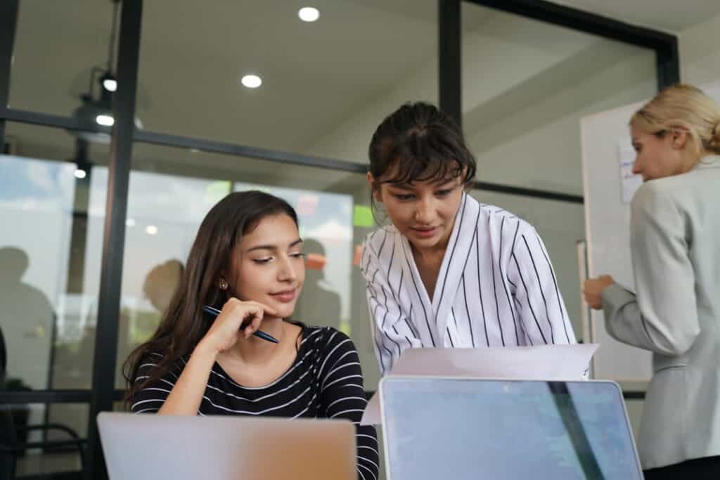 Two people are working in an office. One is seated with a pen, the other standing and looking at papers. Near the whiteboard, another person is engaging with SYNCHRO software, discussing Digital Construction strategies.