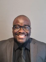A portrait of a smiling black man, Dr. Liu, wearing glasses and a black suit with a black shirt, against a light gray background.