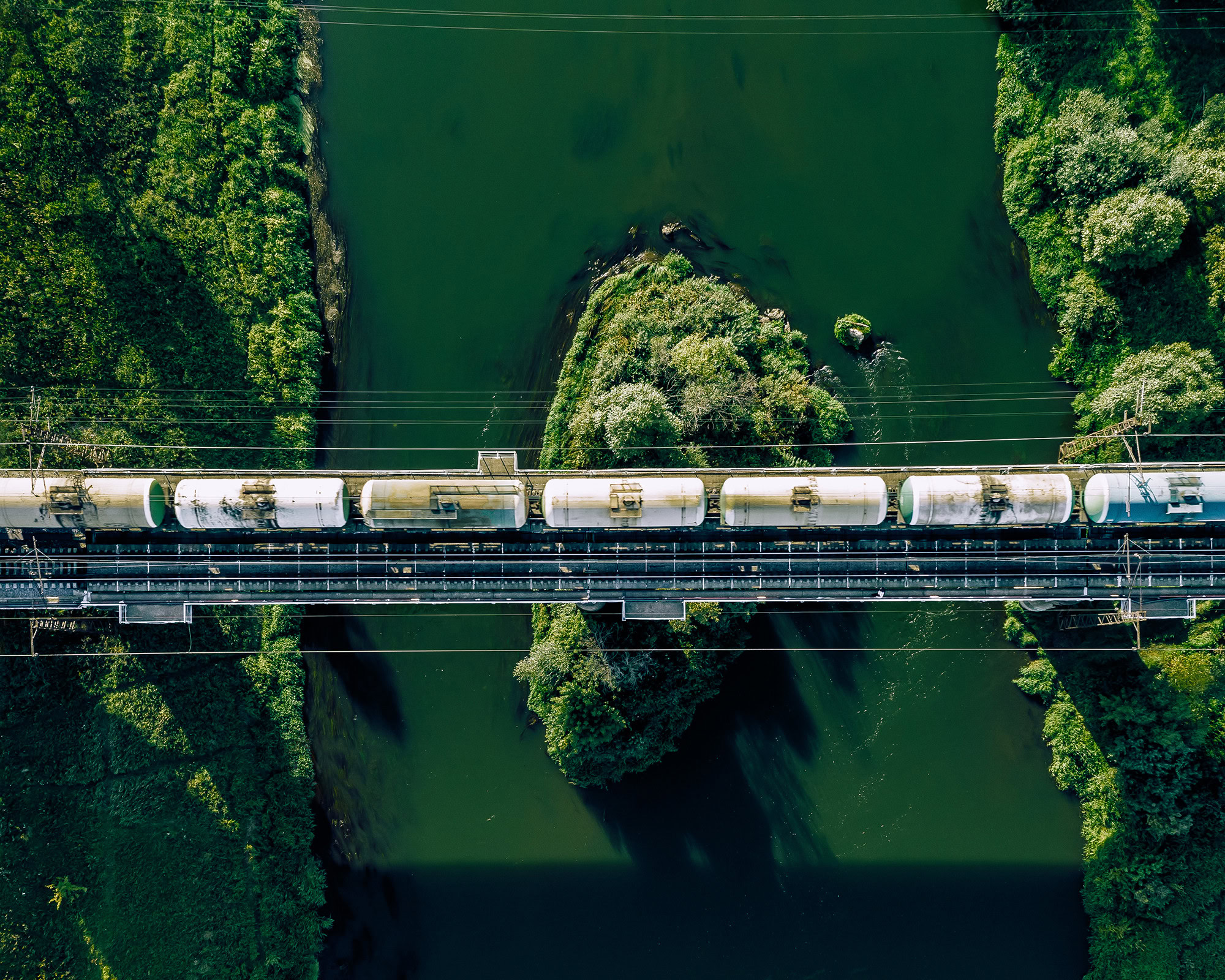 Aerial view of a train crossing a meticulously designed bridge over a green river, surrounded by lush vegetation.