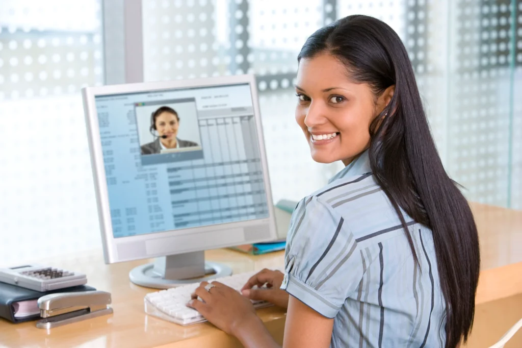 A woman sits at a desk, looking back with a smile while using Analysis Software on her computer, displaying a video call interface.