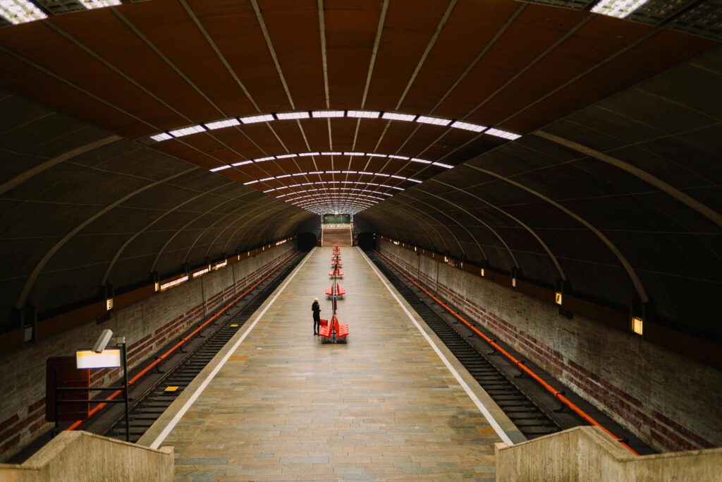A person stands on an empty train platform under an arched roof, with orange cones arranged in a line down the center.