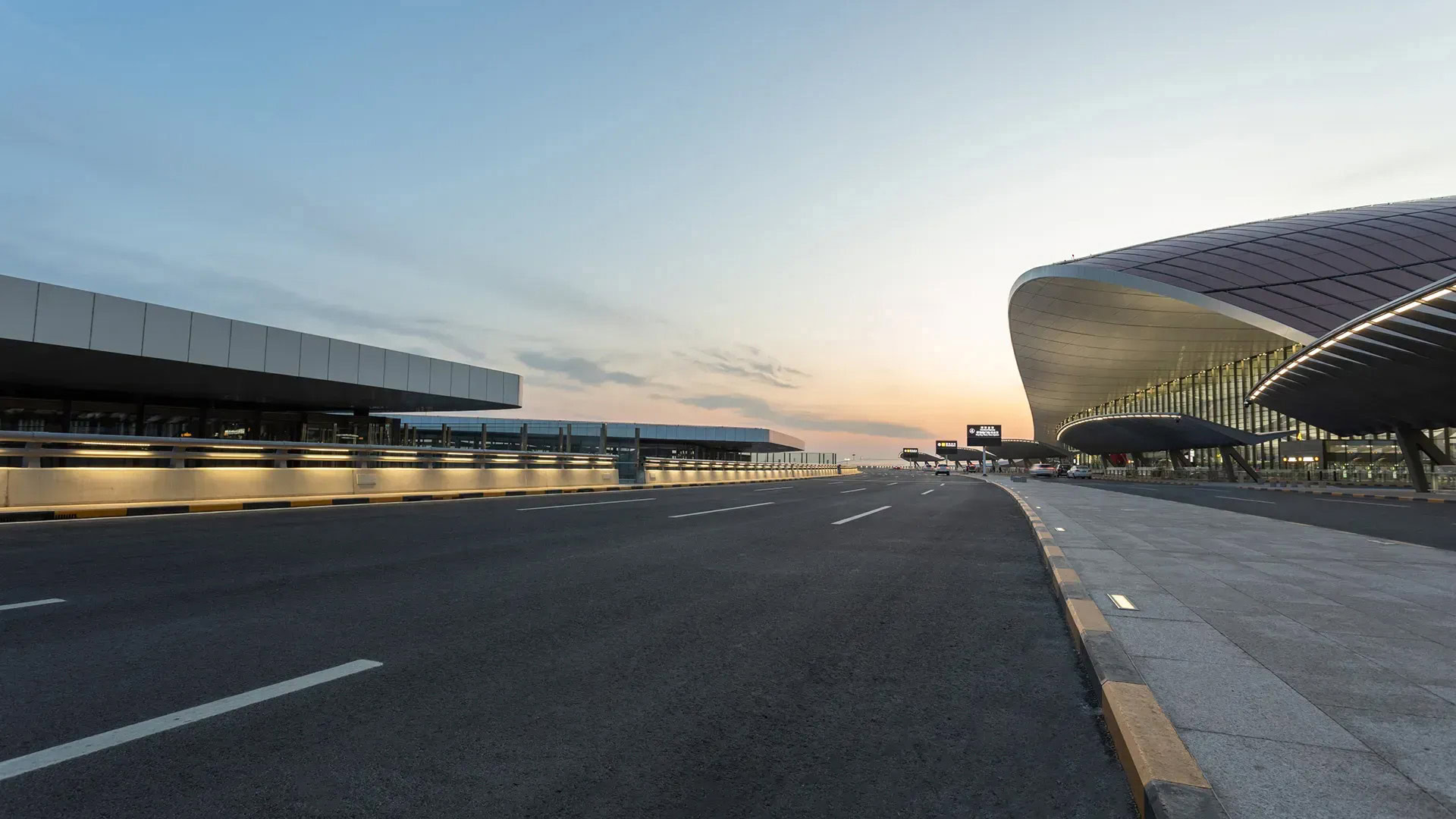 Wide, empty road outside a modern airport terminal at sunset, with glass and metal architecture on the right and clear sky above.