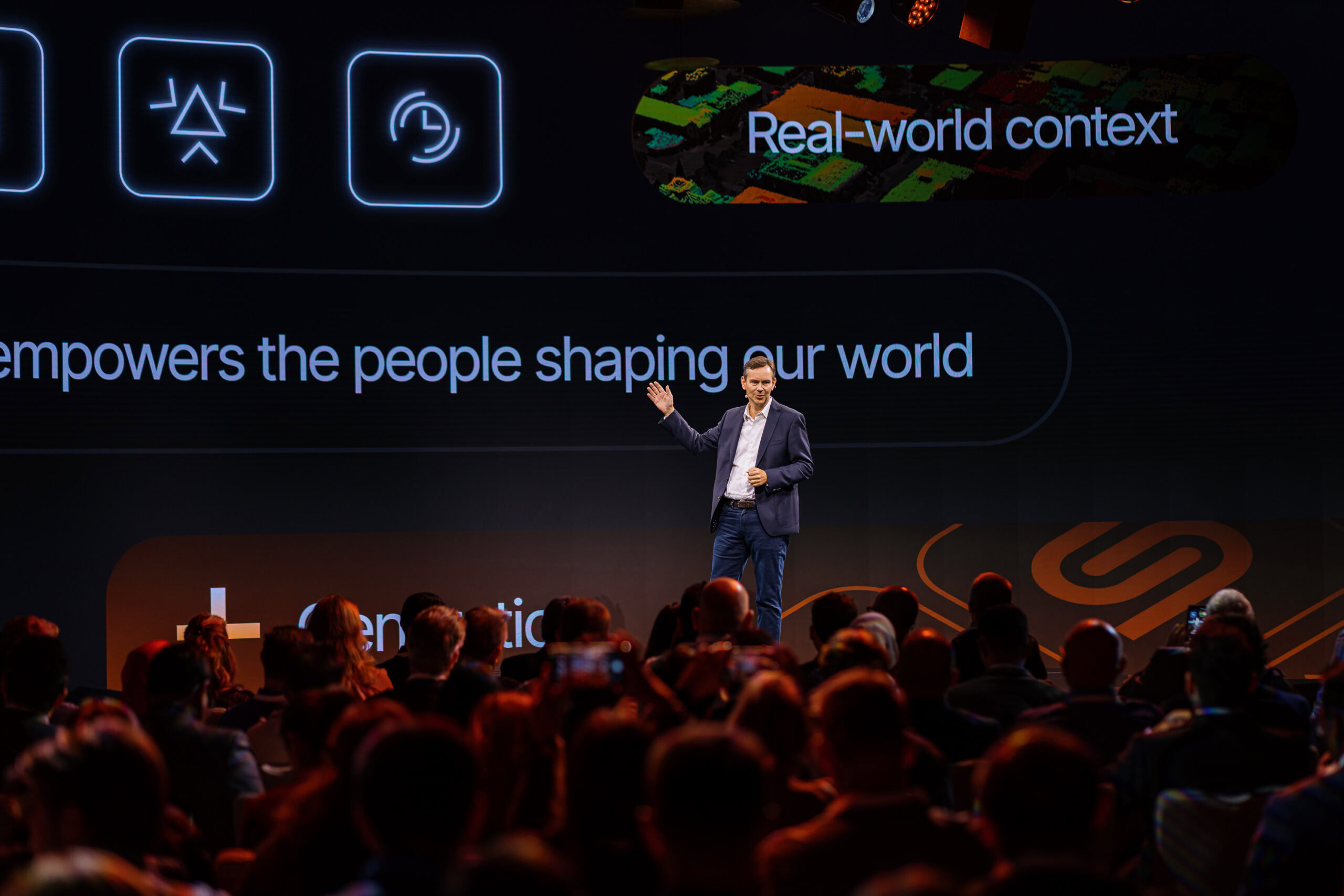 A man in a suit stands on stage giving a presentation to a seated audience, with large digital graphics and text displayed on a screen behind him.
