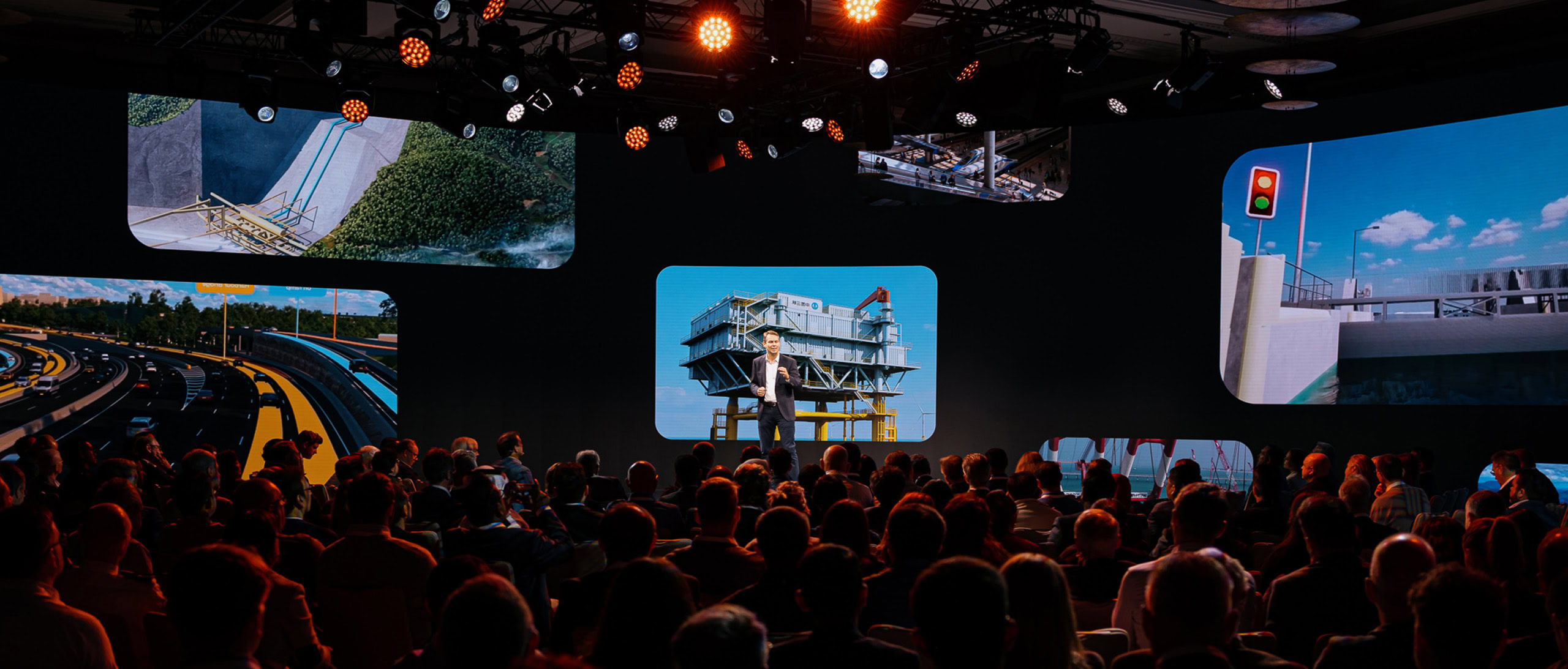A speaker addresses an audience in a dark auditorium with large screens displaying infrastructure and construction images.