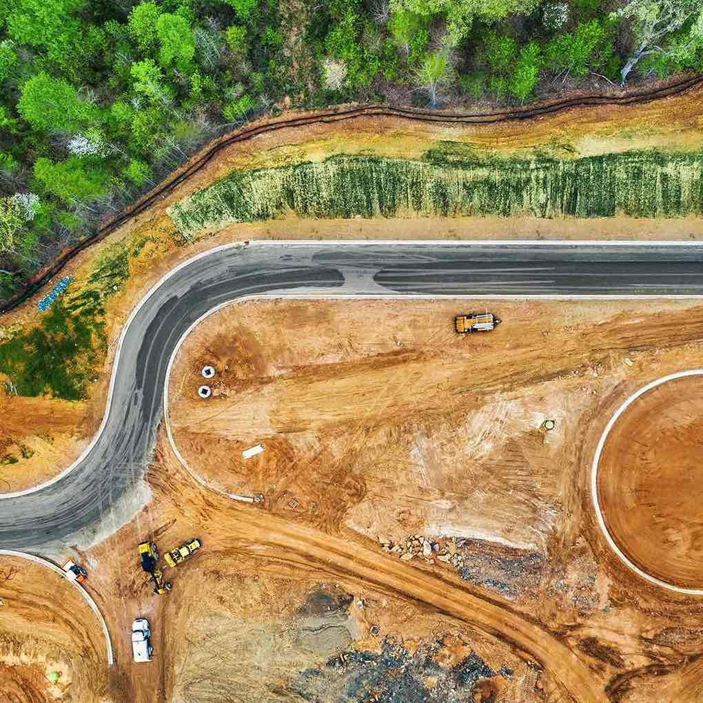Aerial view of a curved road under Highway Construction, bordered by dirt, construction vehicles, and surrounding green trees.
