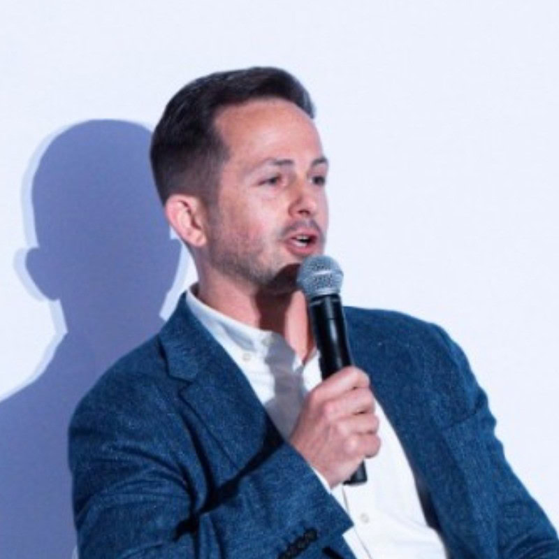 Mężczyzna w niebieskiej marynarce i białej koszuli mówi do ręcznego mikrofonu na jednolitym jasnym tle, reprezentując zespół Bentley.