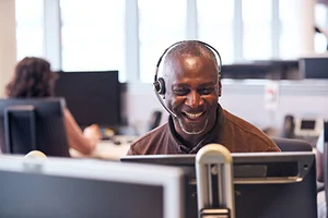 A man wearing a headset smiles while working at a computer in an office environment, possibly providing a user testimonial for OpenSite. Another person sits at a computer in the background.