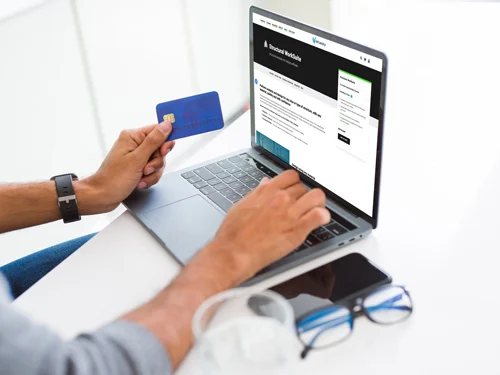 A person shopping online using a laptop, holding a credit card in one hand, with eyeglasses and headphones on the desk next to a Structural WorkSuite manual.