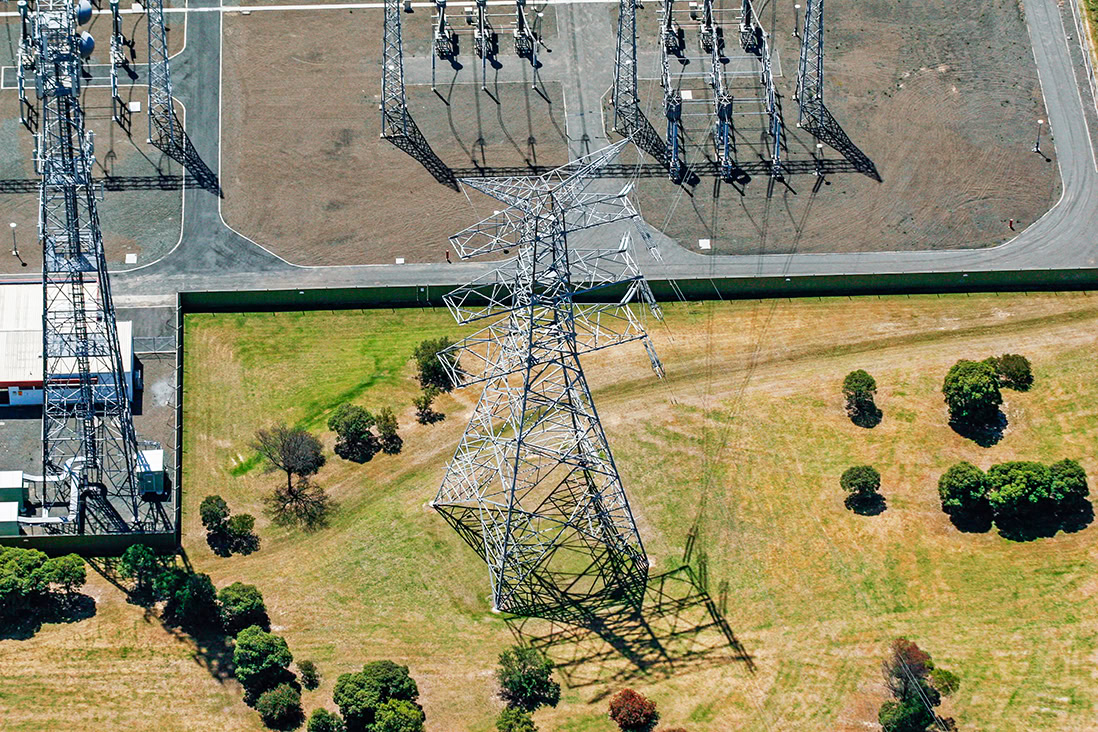 Aerial view of a large electric utilities transmission tower casting a shadow on grass, with nearby trees and a fenced utility area visible.