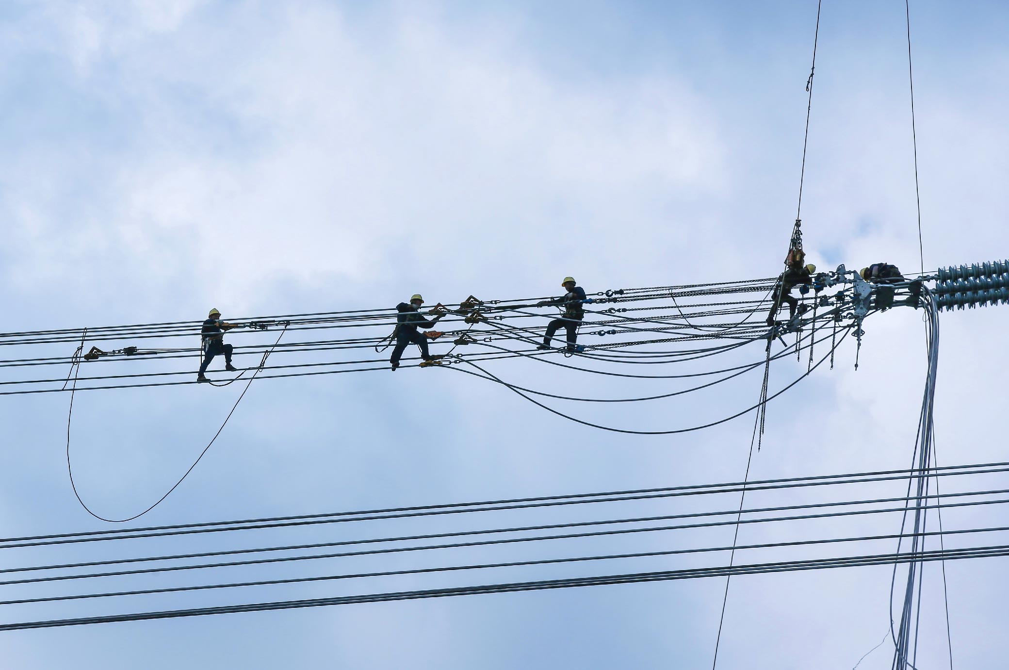 Four workers wearing safety gear are walking and working on high-voltage power lines for an electric utility company against a cloudy sky.