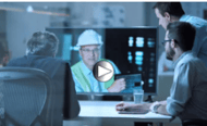 Three men in an office setting are watching an overview video on a large monitor, featuring a person in a white hard hat and safety vest.