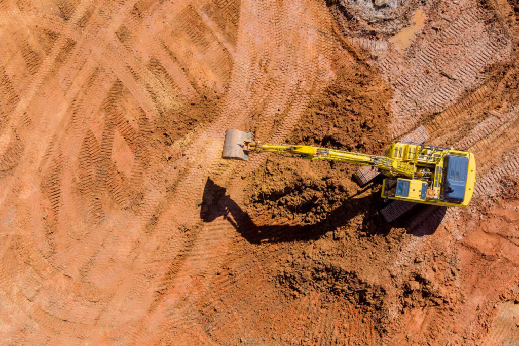 A yellow excavator is digging into reddish-brown soil at a construction site, viewed from above, with visible tire tracks on the ground—showcasing the efficiency of Digital Construction Delivery.