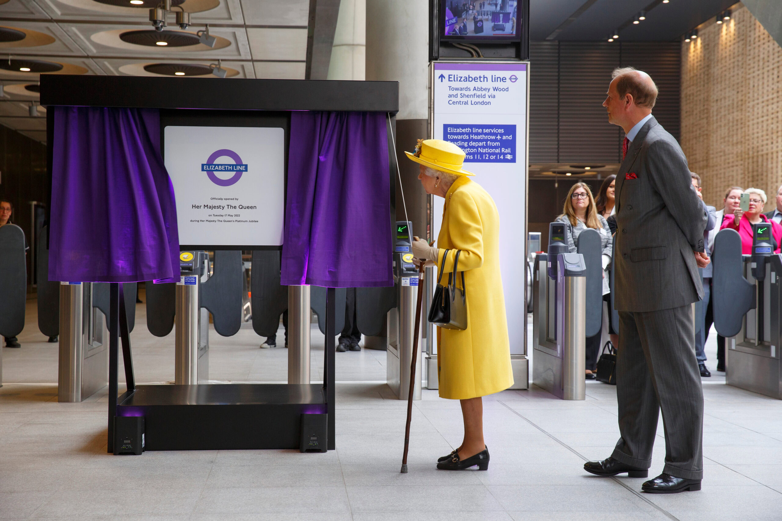 Queen Elizabeth II in a yellow outfit unveils a sign for the Elizabeth line in a train station, with several people observing in the background.
