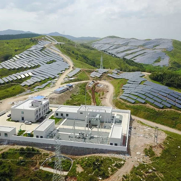 Aerial view of a solar power plant by HUBEI ELECTRIC ENGINEERING, with solar panels covering hillsides and a central building complex surrounded by a fence, set in a green, hilly landscape.