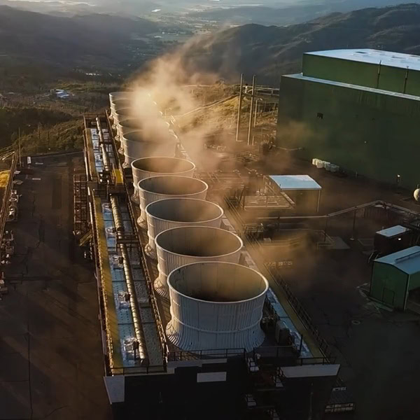 Aerial view of an industrial facility with a row of cooling towers releasing steam, set against a backdrop of hills and scattered buildings.
