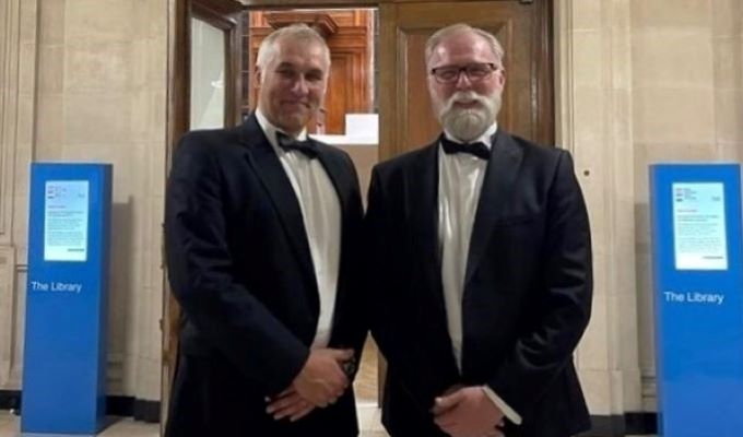 Mark Coates and Rice wear tuxedoes and smiling at the camera while standing in front the library doors