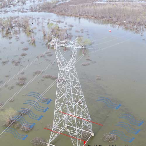 A metal transmission tower stands in a flooded, marshy landscape, with water surrounding its base and partially submerged vegetation visible.