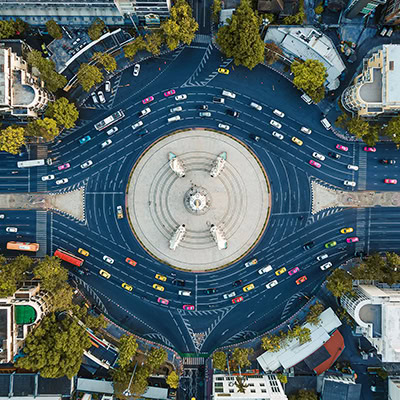 Aerial view of a circular plaza with a central monument, surrounded by roads with moving cars and nearby buildings and trees.