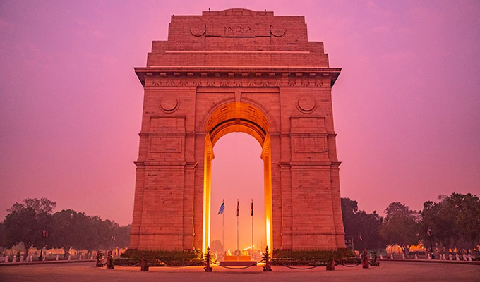 India Gate, an arched monument in New Delhi, is illuminated at dusk as part of the Illuminate India campaign. The sky has a purple hue, and flags are visible at the base of the monument. Trees and pathway are seen in the background, creating a serene setting to say "Thank You" to our heroes.