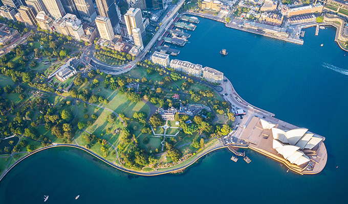 Aerial view of a city waterfront with green parks and distinct architectural structures next to a blue harbor.