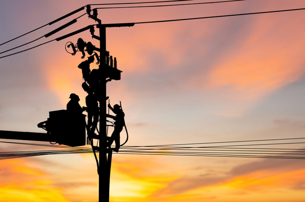 Silhouette of two utility workers repairing electrical wires on a power pole during sunset, with colorful sky in the background.