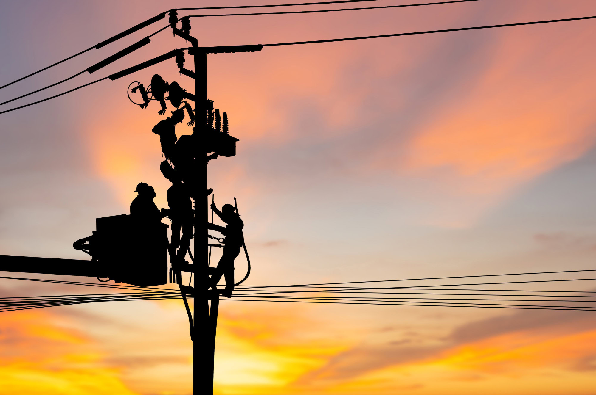 Silhouette of two utility workers repairing electrical wires on a power pole during sunset, with colorful sky in the background.