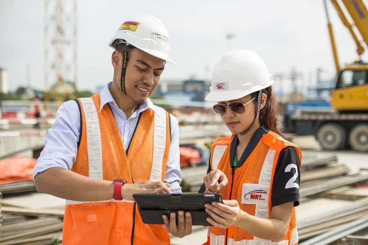 A man and a women wearing hard hats and safety vest at a construction site looking at an iPad.
